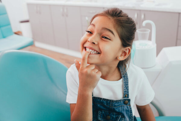 Young girl pointing at teeth, in orthodontist exam chair, smiling, Saxe Orthodontics' children's first orthodontist visit in Summerlin, Las Vegas